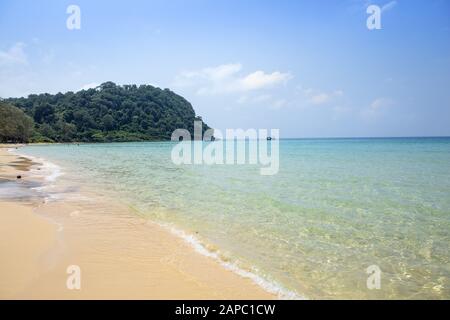 Cambodia, Koh Rong Sanloem. Turquoise waves lapping on a pristine empty beach with a forested cape and wooden fishing boat in the distance Stock Photo