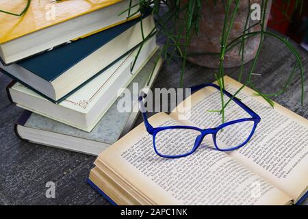 VIERSEN, GERMANY - AUGUST 21. 2019: View on open yellowed book with blue reading glasses and stack of old books on round wood table. Succulent plant b Stock Photo