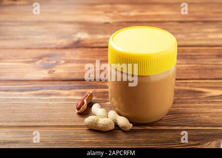 Creamy peanut paste in glass jar with yellow cap and peanuts in the peel scattered on the brown wooden table with copy space for cooking breakfast Stock Photo