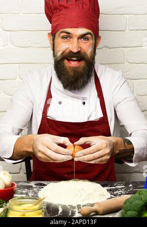 Cook with happy face in burgundy uniform sits by table with flour and kitchenware. Man with beard breaks egg on white brick wall background. Chef makes dough. Cooking process concept. Stock Photo