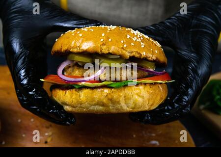 Woman's hands in black rubber gloves are holding juicy bun burger with meat cutlet, lettuce, tomato, shredded cheese and marinated cucumber. Top views Stock Photo