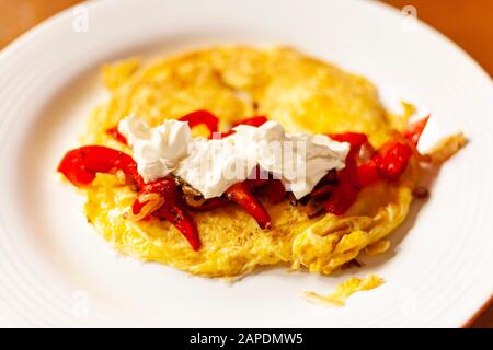 An omelette with sauteed bell peppers, shallots, and sour cream is plated on a white dish with a wooden table as background. Stock Photo