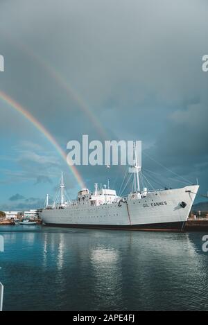VIANA DO CASTELO, PORTUGAL - Jan 03, 2020: A wide shot of a ship by the bay with a rainbow above on a cloudy day Stock Photo