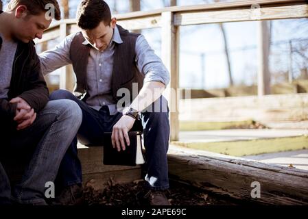 Men sitting on a wooden plank and holding the bible in a garden under sunlight Stock Photo