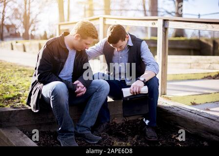 Men sitting on a wooden plank and holding the bible in a garden under sunlight Stock Photo