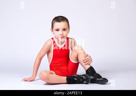 A boy athlete in sportswear and wrestling equipment sits on the floor and looks at the camera cheerfully on a white isolated background. Stock Photo