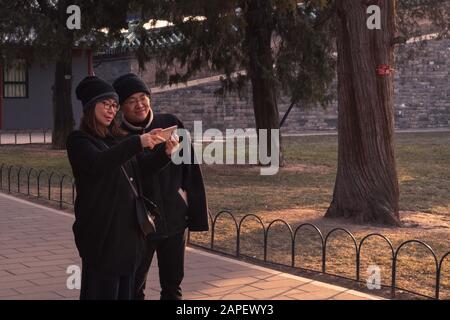 A Chinese couple in a park taking a selfie in Beijing, China, in winter / playing with their phone in a park during Chinese holidays Stock Photo
