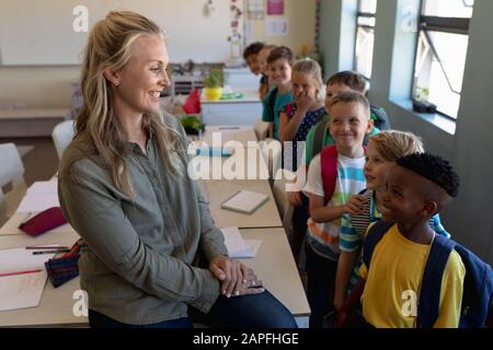 Female school teacher sitting on a desk in a classroom with a group of schoolchildren at elementary Stock Photo