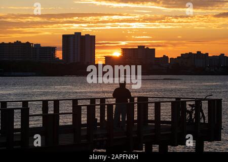A silhouetted man fishing off a pier in the Halifax River in Daytona Bearch, Florida as the sun rises over the skyline of condominiums on the beachfro Stock Photo