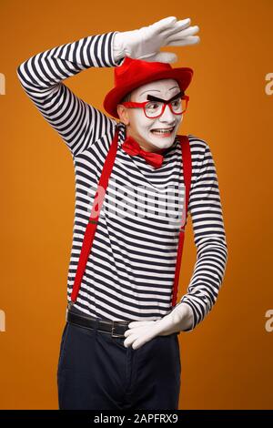 Surprised mime with white face in red hat and striped t-shirt on blank orange background in studio Stock Photo