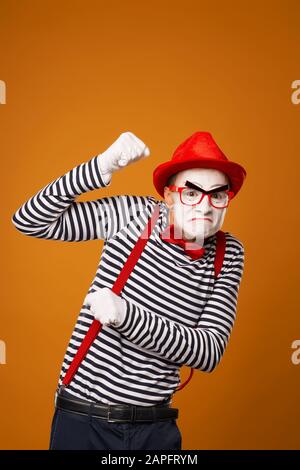 Sad mime with white face in red hat and striped t-shirt on blank orange background in studio Stock Photo