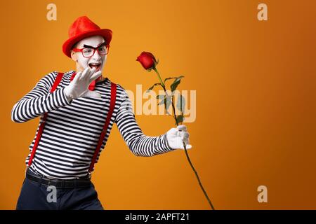 Funny mime man with white face in red hat and striped t-shirt holding rose isolated on empty orange background in studio Stock Photo