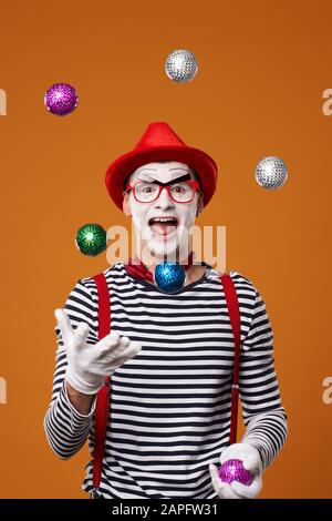 Haappy mime man in vest and red hat juggles with colorful balls on orange empty background Stock Photo