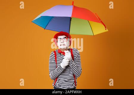 Dissatisfied mime man with multi-colored umbrella isolated on blank orange background in studio Stock Photo