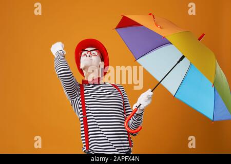 Mime looking up with multi-colored umbrella in hand and showing fist isolated on blank orange background in studio Stock Photo