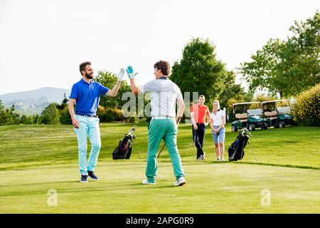 Friends giving high five on golf course, couple watching in background Stock Photo