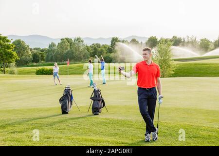 Man taking selfie on golf course, friends playing golf in background Stock Photo