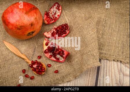 Ripe juicy pomegranate, pieces of pomegranate and pomegranate seeds in a wooden spoon on a background of rough homespun fabric. Close up Stock Photo