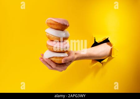Hand offer stack of yummy donuts through a torn hole in yellow paper background Stock Photo