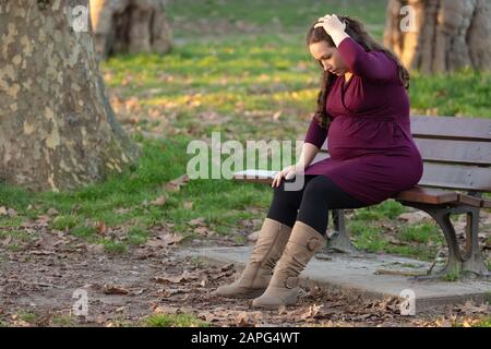 Anxious young pregnant woman on a park bench trying to relax under the warm winter sunshine. Stock Photo