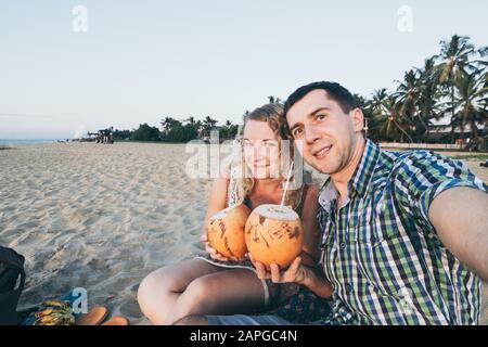 Couple taking selfie at tropical beach in Negombo, Sri Lanka. Holding fresh coconuts in their hands Stock Photo