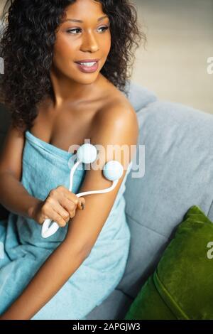 Beauty Day. Woman wearing towel doing her daily skincare routine at home. Sitting on sofa, massaging hands' skin with cosmetic's roller, smiling. Concept of beauty, self-care, cosmetics, youth. Stock Photo