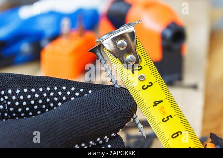 The black screw in hand and is measured by a yellow tape measure with a ruler on a wooden board background. The concept of tools and repair work. Stee Stock Photo