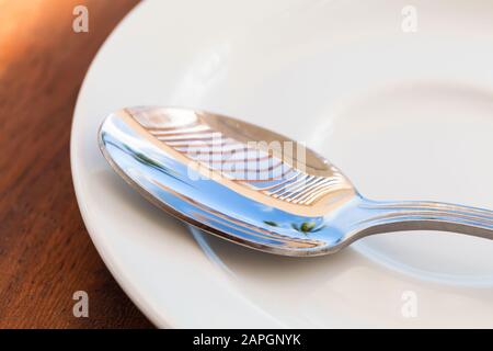 Shiny metal tea spoon with reflections of blue summer sky and palm trees lays on a white ceramic saucer, macro photo with selective focus Stock Photo