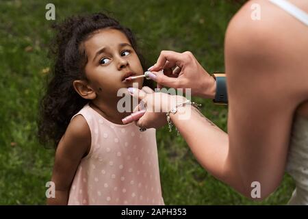 Happy multiethnic family from USA on their European vacation trip. White mother applying hygienic lipstick to her daughter's lips while she is looking