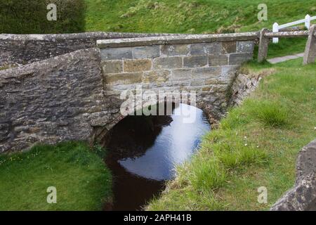 A very small, arched bridge, made from bricks and stone, over a narrow blue stream flowing between high grassy banks. Quaint and cute example of the E