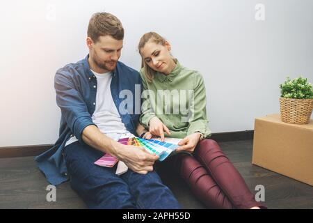 young couple sitting on the floor in the room and choosing paint color from swatch for new interior design Stock Photo