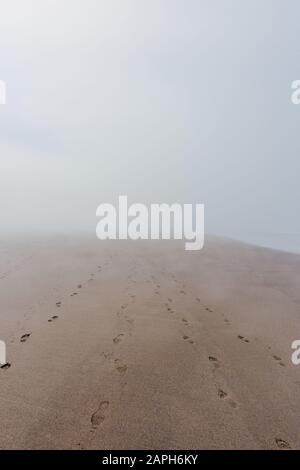 Ocean beach in dense fog with footprints on sand leading nowhere Stock Photo