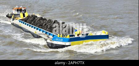 Close up long Hercules barge & load of soil from boring of Thames Tideway Tunnel sewer project tug boat pushing bow wave down river London England UK Stock Photo