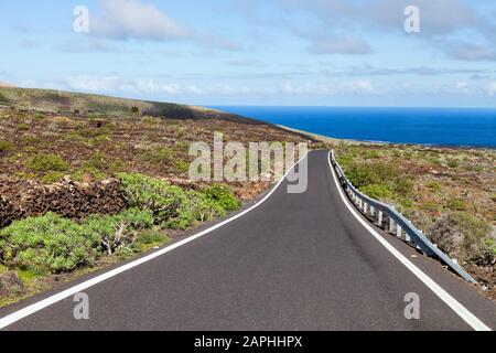 Empty asphalt road through hilly volcanic countryside with green plants, towards Atlantic Ocean coast, Lanzarote, Canary Islands, Spain . Stock Photo