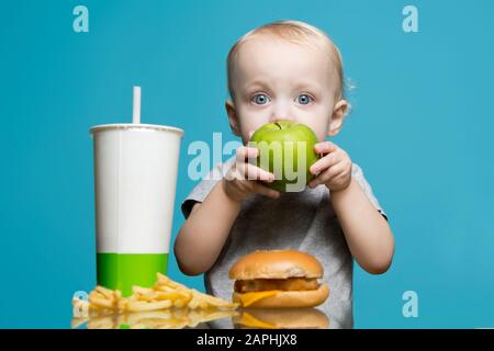 little girl eating an apple, while on the table there was a harmful burger, soda and french fries Stock Photo