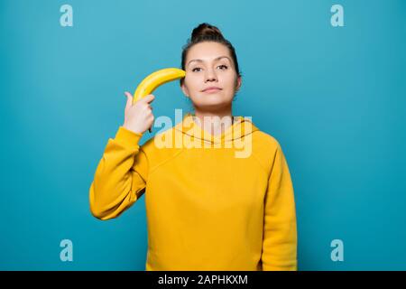 girl in a bright jacket holds a banana near her head in the manner of a revolver Stock Photo