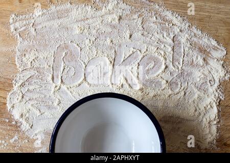 The word 'Bake!' handwritten in scattered flour on a wooden chopping board with empty enamel bowl below. Overhead shot. Stock Photo