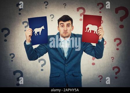 Undecided young man holding two brochures has to choose between donkeys and elephants. Perplexed and confused person with question marks around head. Stock Photo