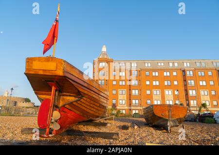 Stern view of two classic old-fashioned boats pulled up on stony beach with large red brick apartment building in background glowing in light from sun Stock Photo
