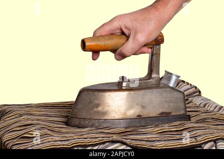 Vintage ironing tool household. Hand of a man, hold an old rusty professional electric tailor iron at the wooden handle in front of yellow background. Stock Photo