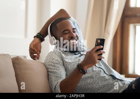 African guy in headphones resting on couch using smartphone laughing Stock Photo