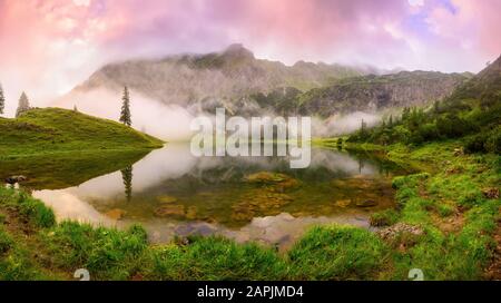 Lake surrounded by mountains at dawn, with colorful clouds and wafts of mist reflected in the water Stock Photo