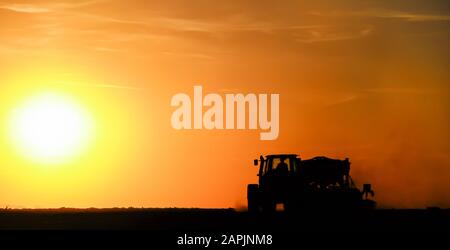 Silhouette of a tractor sowing seeds in a field in a cloud of dust against the background of the setting sun. Stock Photo