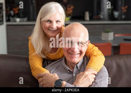 Cheerful, romantic senior couple hugging while sitting on sofa at home Stock Photo