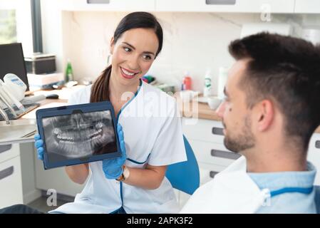 Female dentist showing teeth x-ray on digital tablet screen. Patient sitting on chair in professional dental clinic. Stock Photo