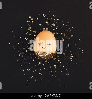 Creative easter egg with cute face and sleepy eyes on black background with golden glitter confetti. Stock Photo