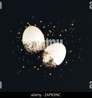White easter eggs in golden glitter confetti and stars on black background. Stock Photo