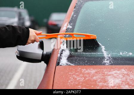 Brush in mans hand. Man clears snow from icy windshield of car. Cleaning orange automobile window. Stock Photo