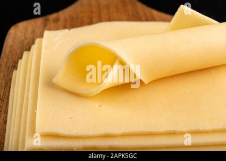 Slices of yellow cheese on a wooden board. Food from the market on the kitchen table. Dark background. Stock Photo