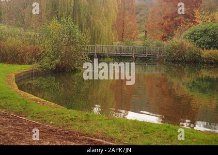 A view across a corner of a lake to a wooden footbridge with trees in the background showing their autumn colours and reflected in the water Stock Photo